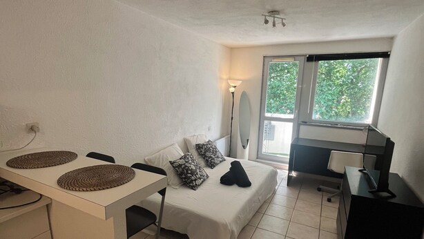 A small, well-lit room with a bed, bedside table, and a desk with a chair. There is a window providing natural light and a view of greenery outside. - Studio in the center of Toulouse, 7 minutes by bike from Place Saint Pierre (Toulouse)