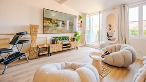 A modern living room with a large painting, a wooden cabinet, a black chair, a beige pouf, and a window with curtains. - Le 6ème Sens: a getaway for two in Toulon (Toulon)