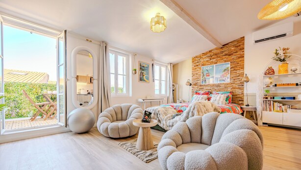 A modern living room with a large window, a round seating area, a bed with a colorful blanket, and a brick accent wall. - Le 6ème Sens: a getaway for two in Toulon (Toulon)