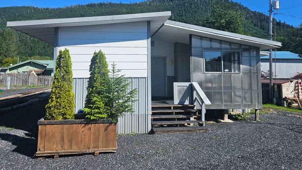 A small, single-story building with a porch and a wooden planter with a coniferous tree.