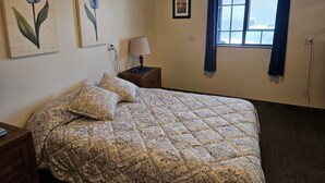 A bedroom with a bed, two wall-mounted paintings, a nightstand with a lamp, a window with curtains, and a small picture on the wall.