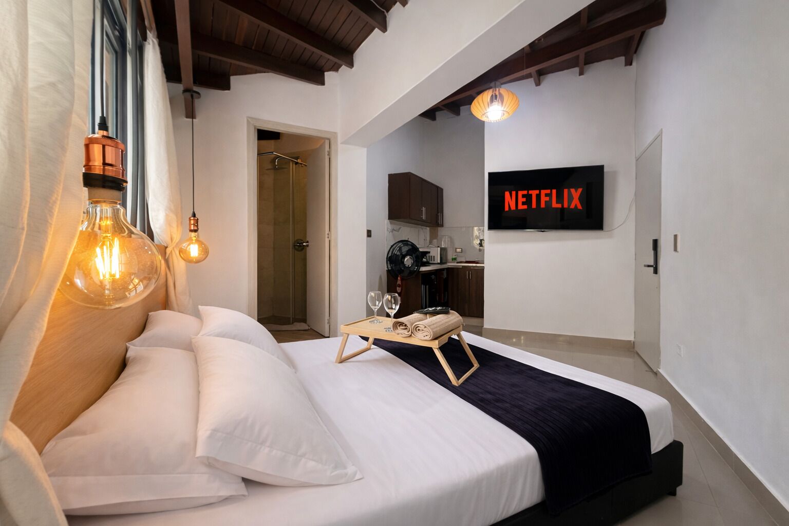 Une chambre d’hôtel moderne équipée d’un lit, de lampes de chevet, d’un plateau avec des verres et des serviettes, d’un coin cuisine et d’un téléviseur fixé au mur affichant « NETFLIX ».