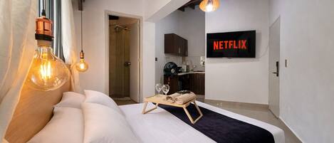 Une chambre d’hôtel moderne équipée d’un lit, de lampes de chevet, d’un plateau avec des verres et des serviettes, d’un coin cuisine et d’un téléviseur fixé au mur affichant « NETFLIX ».