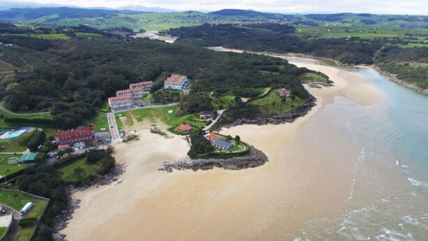 An aerial view of a coastal area with a sandy beach, a river, and a cluster of buildings with red roofs. - Front line duplex - La Arena 4607 (Arnuero)