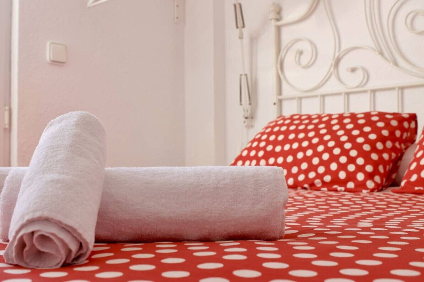 Una cama bien tendida con toallas enrolladas, una colcha roja y blanca con lunares y un cabecero de metal blanco.