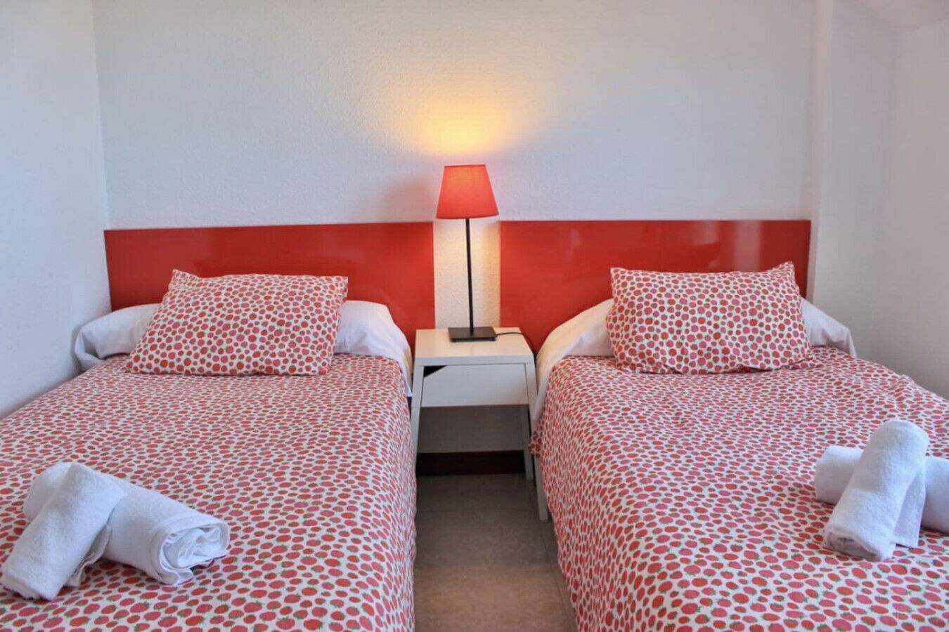 A hotel room with two beds, each with a red and white polka dot bedspread, a white nightstand with a lamp, and towels on the beds.