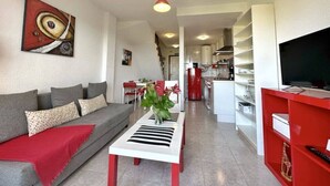 A modern living room with a sofa, a coffee table with a plant, a red TV stand, and a kitchen area in the background. - Front line duplex - La Arena 4607 (Arnuero)