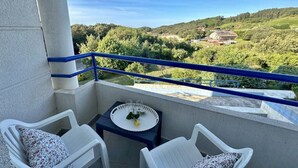 A balcony with white chairs, a small table with a tray, and a view of green hills and a building. - Front line duplex - La Arena 4607 (Arnuero)