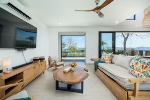 Ein modernes Wohnzimmer mit Flachbildfernseher, Holzmöbeln, Deckenventilator und großen Fenstern mit Blick ins Freie.