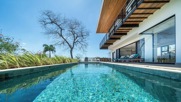 Une piscine à l’eau claire, entourée d’un bâtiment moderne doté de grandes fenêtres et d’une terrasse en bois.