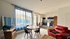 Uma sala de estar com televisão, uma mesa de jantar, cadeiras, um sofá vermelho e uma cozinha ao fundo.