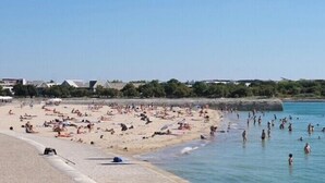 Uma praia com pessoas tomando sol e nadando, uma faixa de areia e edifícios ao fundo.