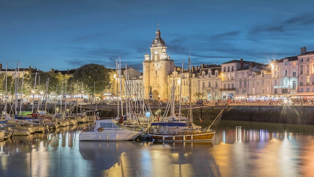 Uma marina com barcos atracados, um edifício imponente com torre de relógio e um calçadão tranquilo.