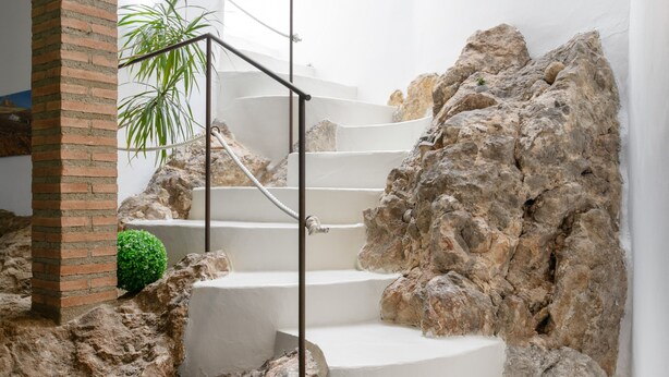 Una scala con gradini bianchi che conduce a un pianerottolo, circondata da un muro di roccia e una piccola pianta in vaso.