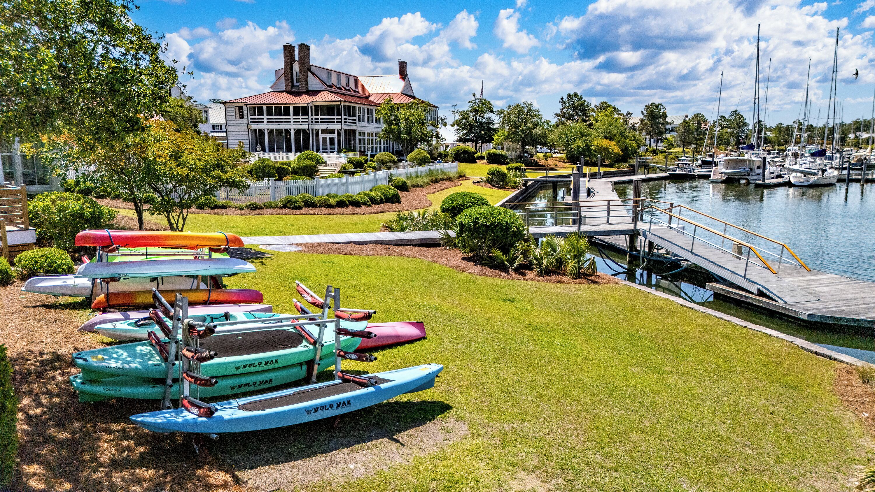A marina with boats docked, a grassy area with a building in the background, and a clear sky.