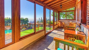 A covered patio with large windows, wooden furniture, and a view of a garden. - Casa El Algarrobo – Home above the clouds with sea views (Puntagorda)