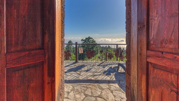 An open wooden door leading to a stone-paved area with a metal fence and a clear sky. - Casa El Algarrobo – Home above the clouds with sea views (Puntagorda)