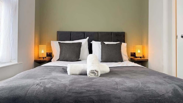 Cama bem arrumada com colcha cinza, duas almofadas e uma toalha enrolada. Existem duas luminárias de cabeceira em cada lado da cama.