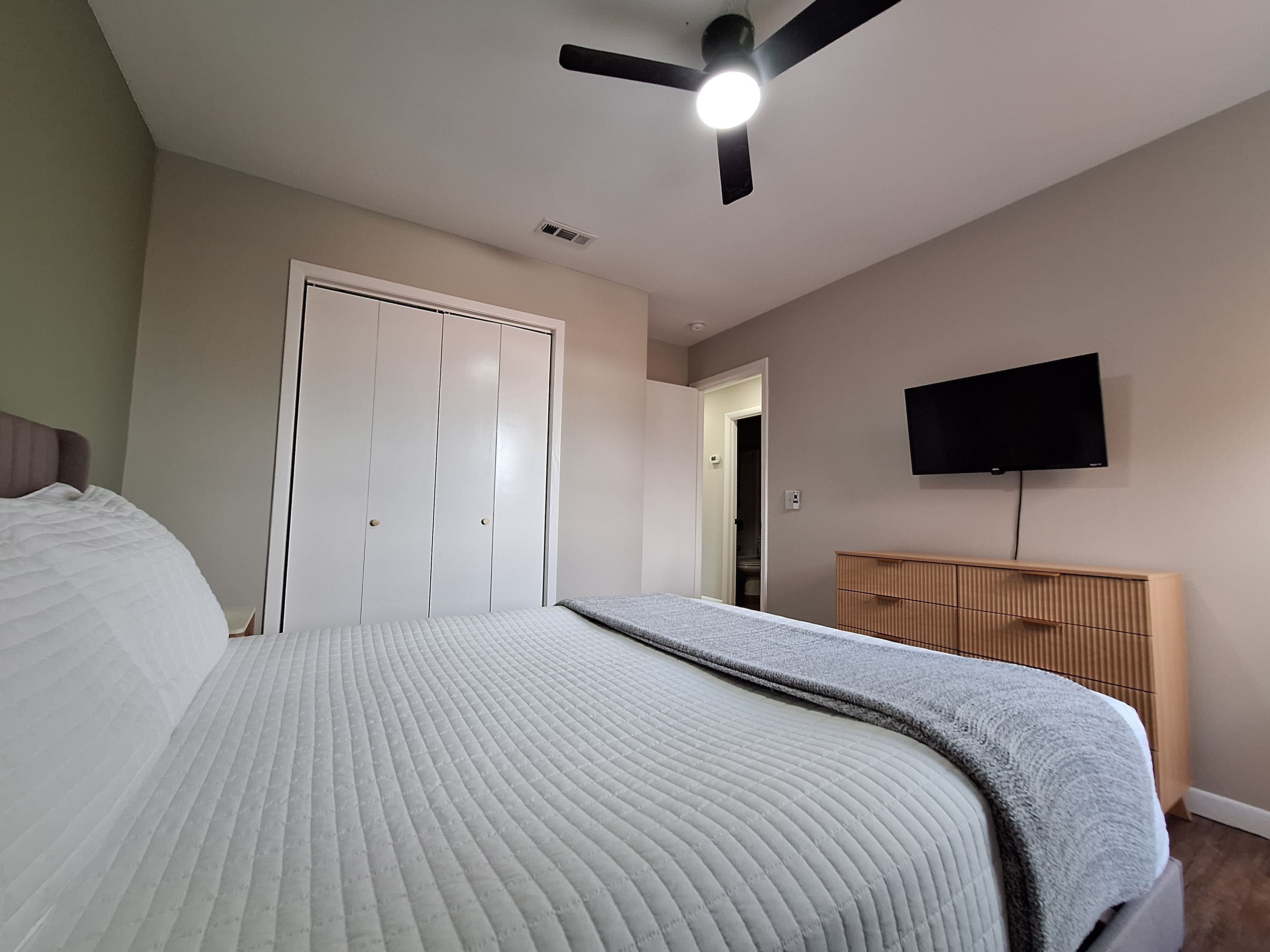 A bedroom with a bed, a ceiling fan, a wardrobe, a television, and a door leading to another room.