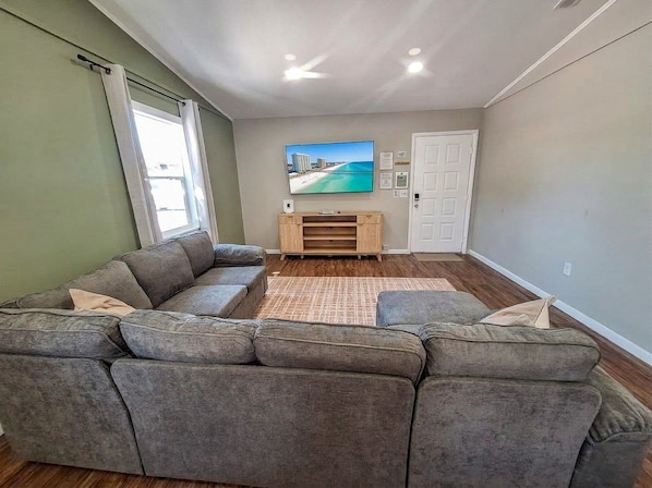 Un salon comprenant un canapé d’angle, un téléviseur à écran plat fixé au mur, un meuble de rangement en bois et une porte.
