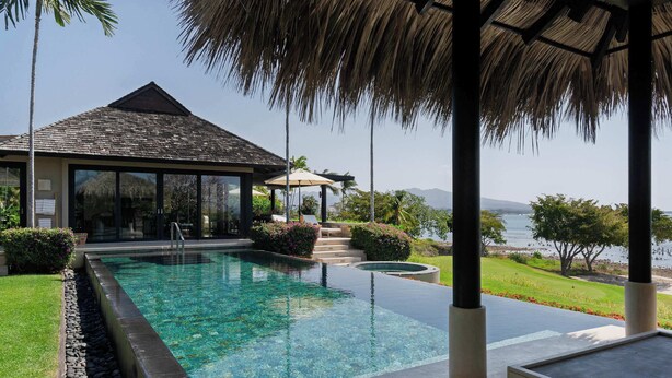Uma piscina com água cristalina, uma construção com telhado de palha e vista para a água.