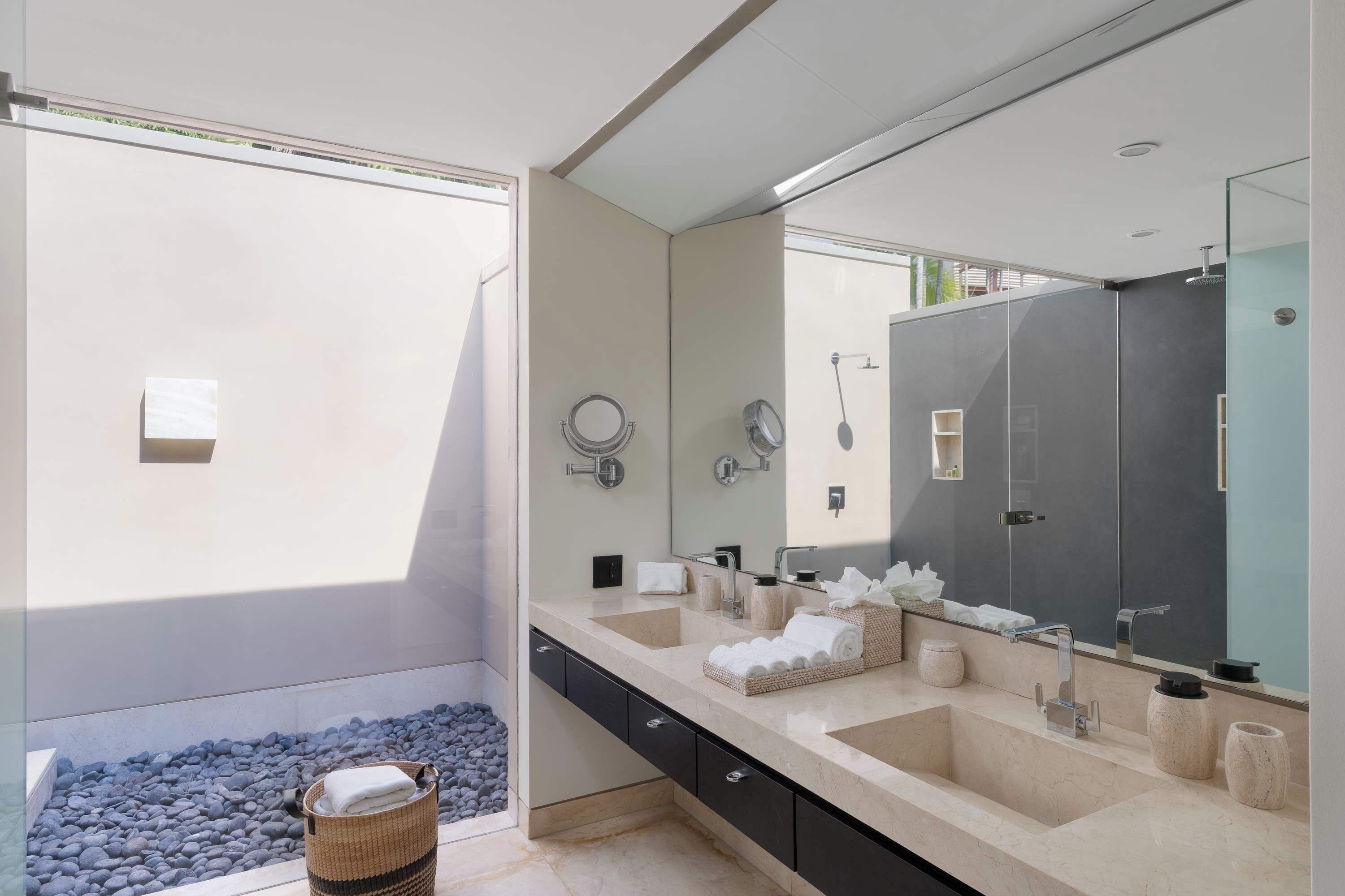 A modern bathroom with a large mirror, two sinks, a shower, and a small seating area with a basket.