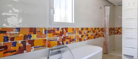 A modern bathroom with a large bathtub, a window, and colorful mosaic tiles on the wall.