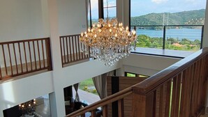 Un interior espacioso con una escalera de madera, un candelabro y amplios ventanales que ofrecen vistas a un lago.