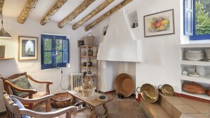 A rustic room with exposed wooden beams, a blue-framed window, a shelf with plates, and a wall-mounted painting.