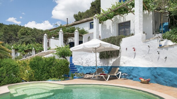 A pool area with lounge chairs and an umbrella, surrounded by a white building with blue accents and greenery.