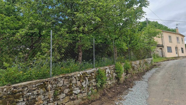 A rural road with a stone wall and a fence, a house with a chimney, and lush green trees.