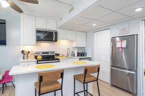 A modern kitchen with white cabinets, a stainless steel refrigerator, a microwave, and a stove. There are two bar stools and a pink chair. - Walk-Out to Sand - Oceanfront Condo (Ocean City)