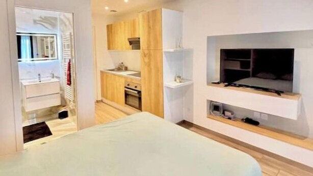 Un dormitorio moderno con cama, televisor montado en la pared y una kitchenette al fondo.