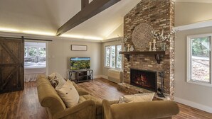A living room with a fireplace, wooden floors, a sofa, and a television. - Serenity on Dogwood is nestled within a wooded section of Hound Ears community. (Boone)
