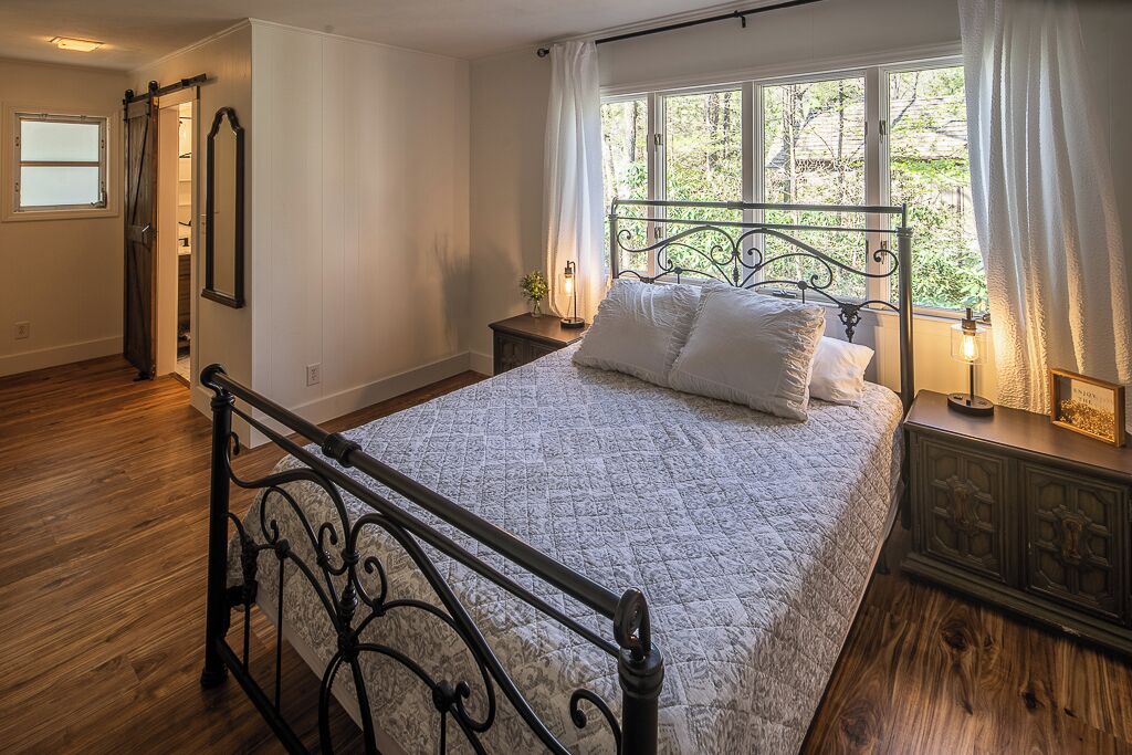 A bedroom with a bed, wooden flooring, a window with curtains, a nightstand, and a wall-mounted mirror.