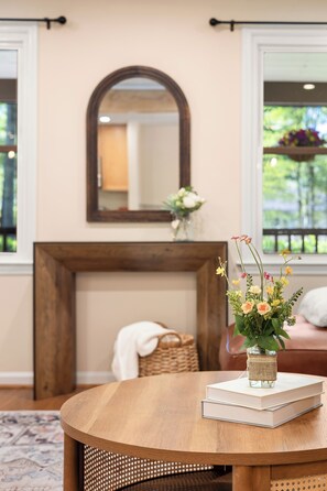 Ein runder Holztisch mit Blumen in einer Vase und Büchern, ein Spiegel über einem Kamin, und ein Korbstuhl.