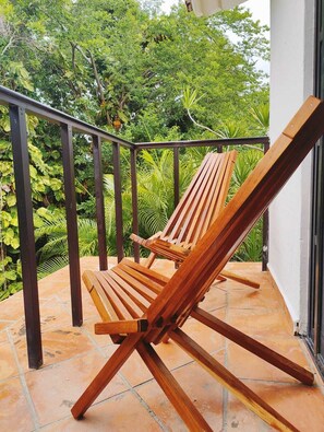 A wooden lounge chair on a tiled patio with a metal railing, surrounded by lush greenery. - Villa in Playacar with a view of the golf course—5-minute walk to the beach and 5th Avenue (Playa del Carmen)