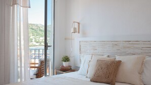 A bedroom with a large bed, a bedside table with a plant, a chair, and a view of the outdoors.