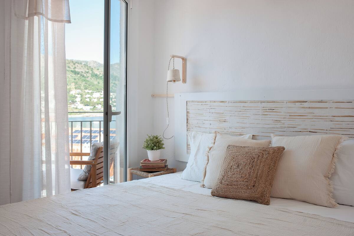 A bedroom with a large bed, a bedside table with a plant, a chair, and a view of the outdoors.