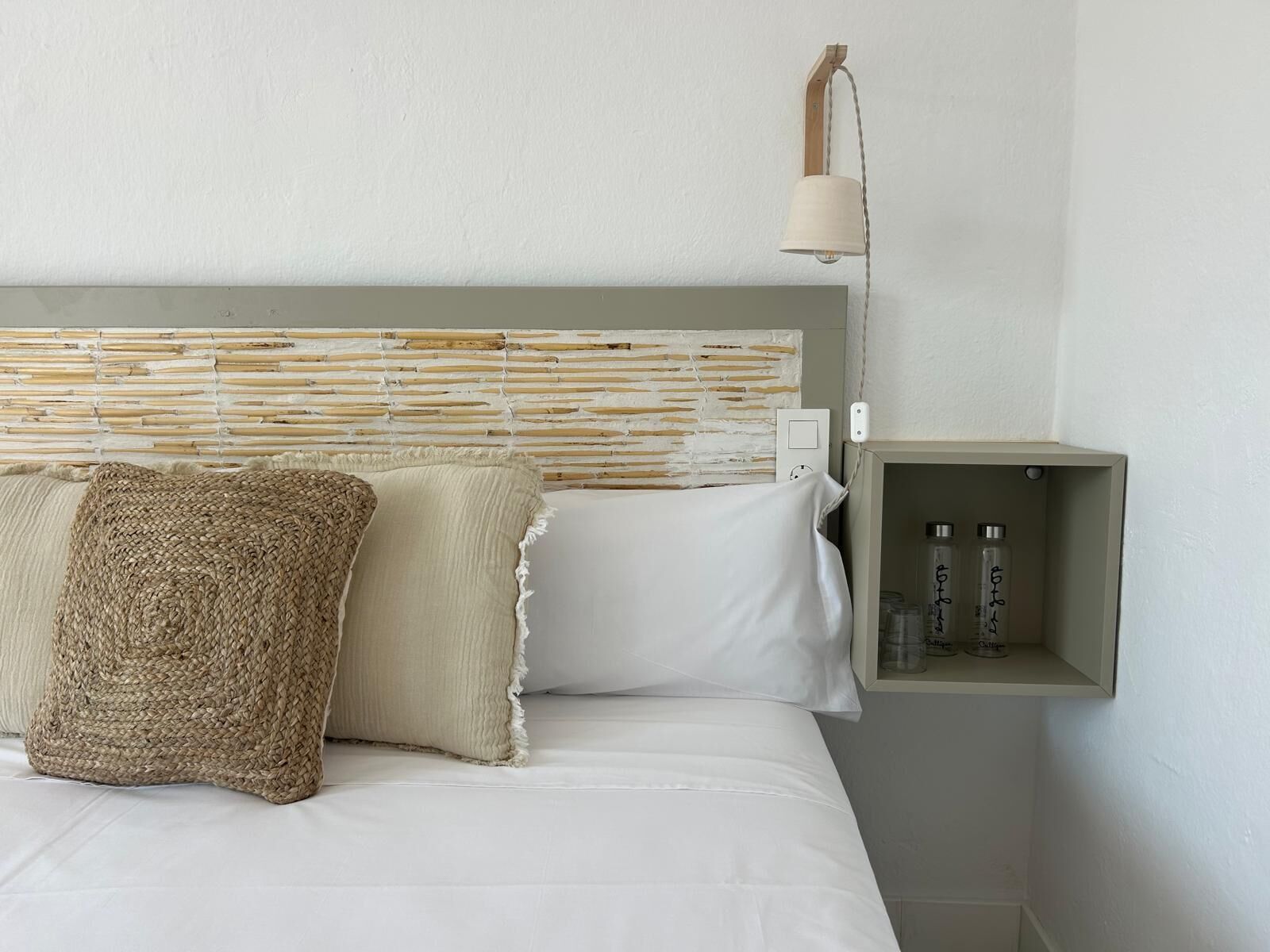 Una cama con ropa de cama blanca, un cojín de tejido y un cojín sencillo. Se ve una lámpara de pared y un pequeño estante con botellas.