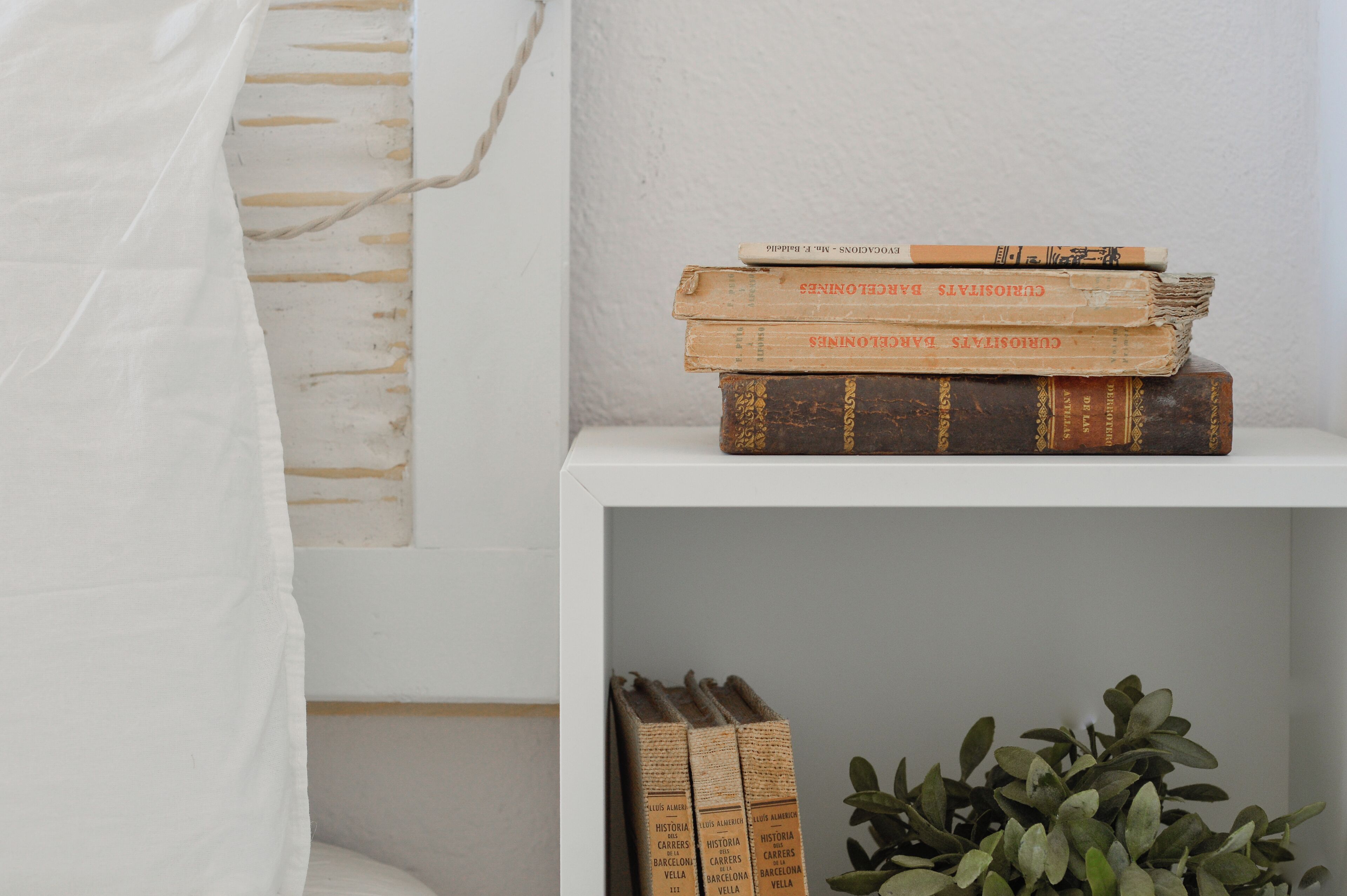Eine Stapel Bücher auf einem weißen Regal, ein weißer Vorhang und eine Pflanze.