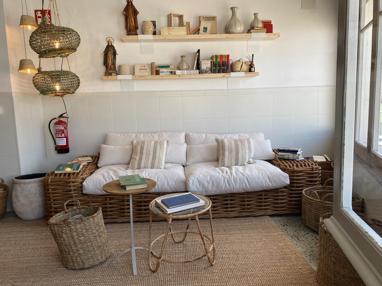 Una sala de estar acogedora con un sofá blanco, cestas de mimbre, una pequeña mesa redonda y estanterías con libros y objetos decorativos.