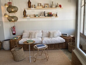A cozy living room with a white sofa, wicker baskets, a small round table, and shelves with books and decorative items. - Hostal Boutique Es Grop (Port de la Selva)