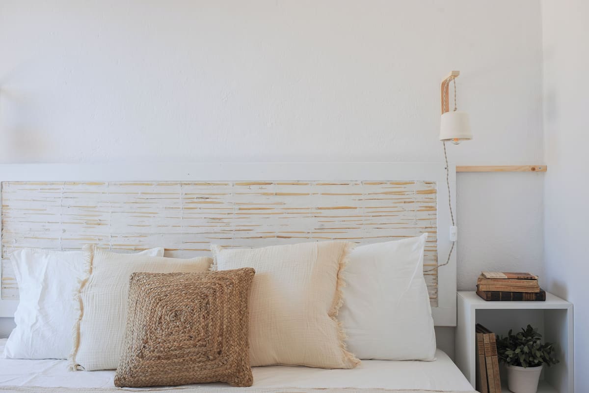 A bed with white bedding and a woven throw pillow, a bedside table with a lamp, and a shelf with books and a plant.