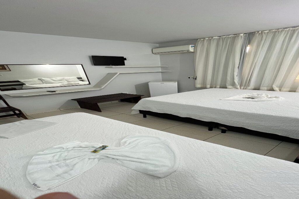 A hotel room with two beds, a flat-screen TV, an air conditioner, and a large window with curtains.