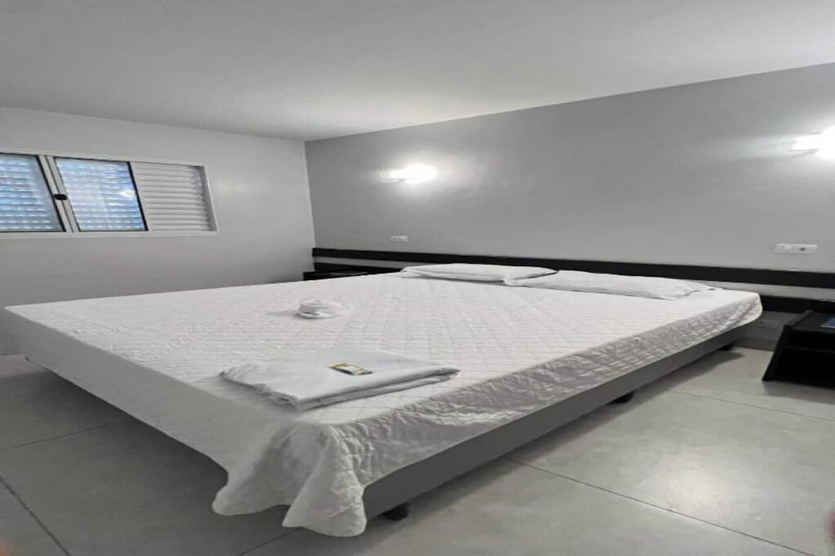 Quarto de hotel com uma cama grande, uma mesinha de cabeceira, uma janela com persianas e uma luminária na parede.
