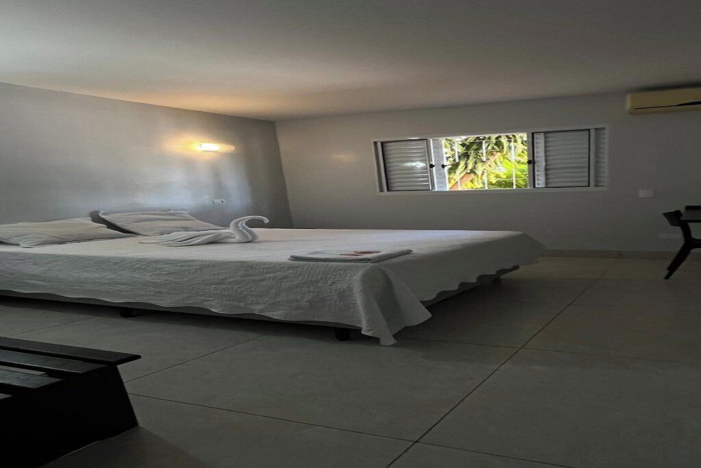 A bedroom with a bed, a bedside table, a window with blinds, and a wall-mounted air conditioner.