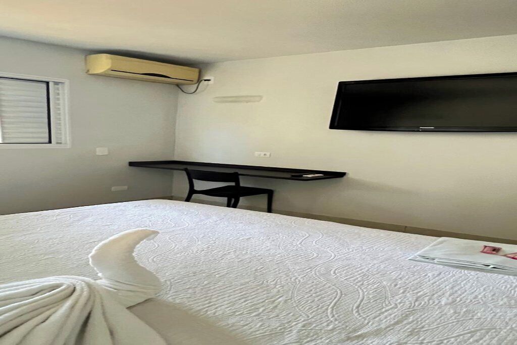 A hotel room with a bed, a desk, a chair, a flat-screen TV mounted on the wall, and an air conditioning unit.