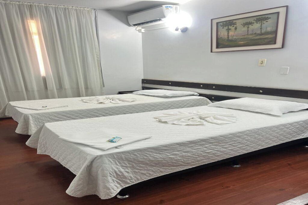 A hotel room with two beds, a painting on the wall, an air conditioning unit, and a window with curtains.