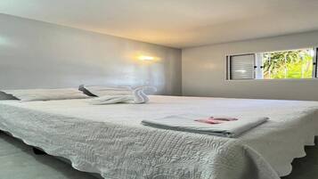 A neatly made bed with a swan pillow and a folded towel, a wall-mounted light, and a window with blinds.